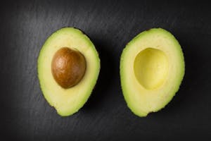 A halved avocado on a dark surface. The left half contains the brown seed, while the right half shows the hollow indent where it was. The bright green flesh of this fruit contrasts against the dark background.